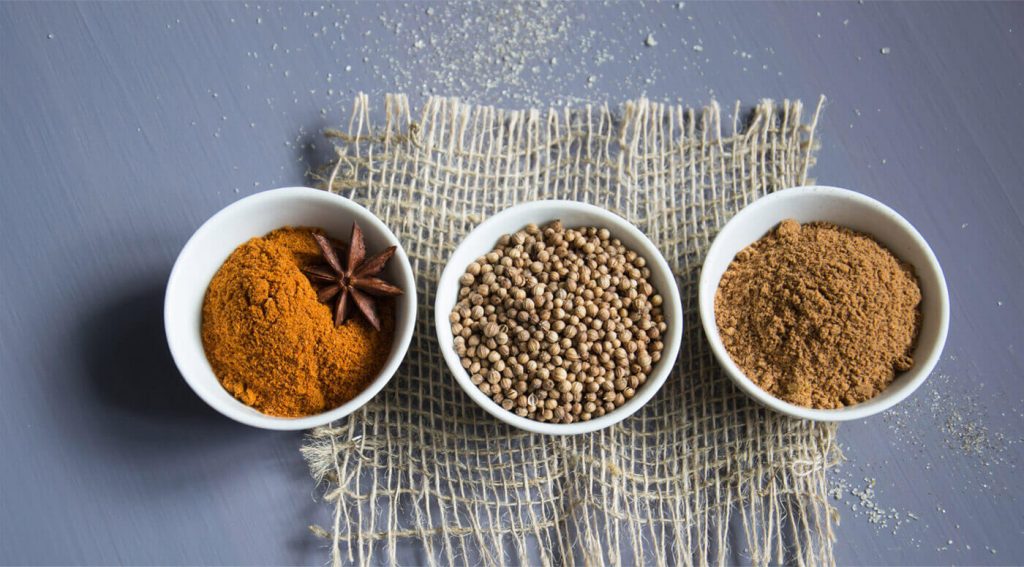 Three white bowls filled with spices are arranged in a row on a rustic woven mat over a grey surface. The left bowl contains a red-orange spice with star anise, the middle bowl holds whole coriander seeds, and the right bowl has a brown spice powder.
