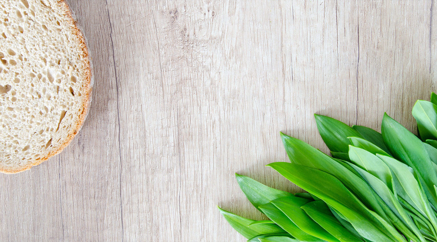 A slice of bread lies on the left side of a light wooden surface. On the right side, there's a bunch of fresh green leaves. The composition suggests ingredients for a healthy meal or sandwich.