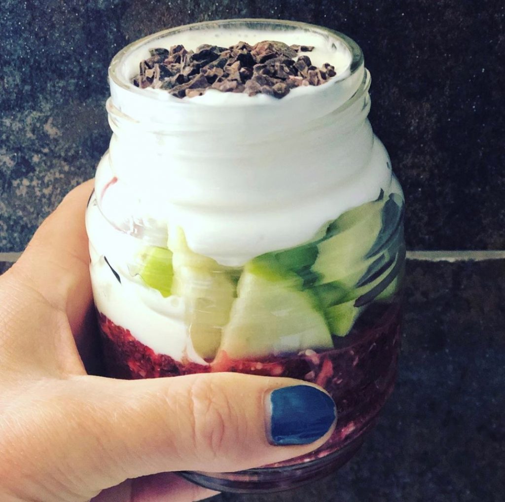 A hand with blue-painted nails holds a glass jar layered with yogurt, apple slices, berry compote, and topped with chocolate shavings. The jar is positioned against a dark, textured background.